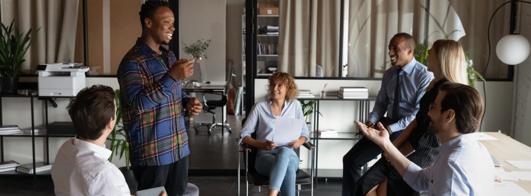 Smiling African American male leader talk hold meeting with diverse colleagues coworkers at workplace. Happy multiethnic businesspeople speak discuss business ideas at team meeting in office.
